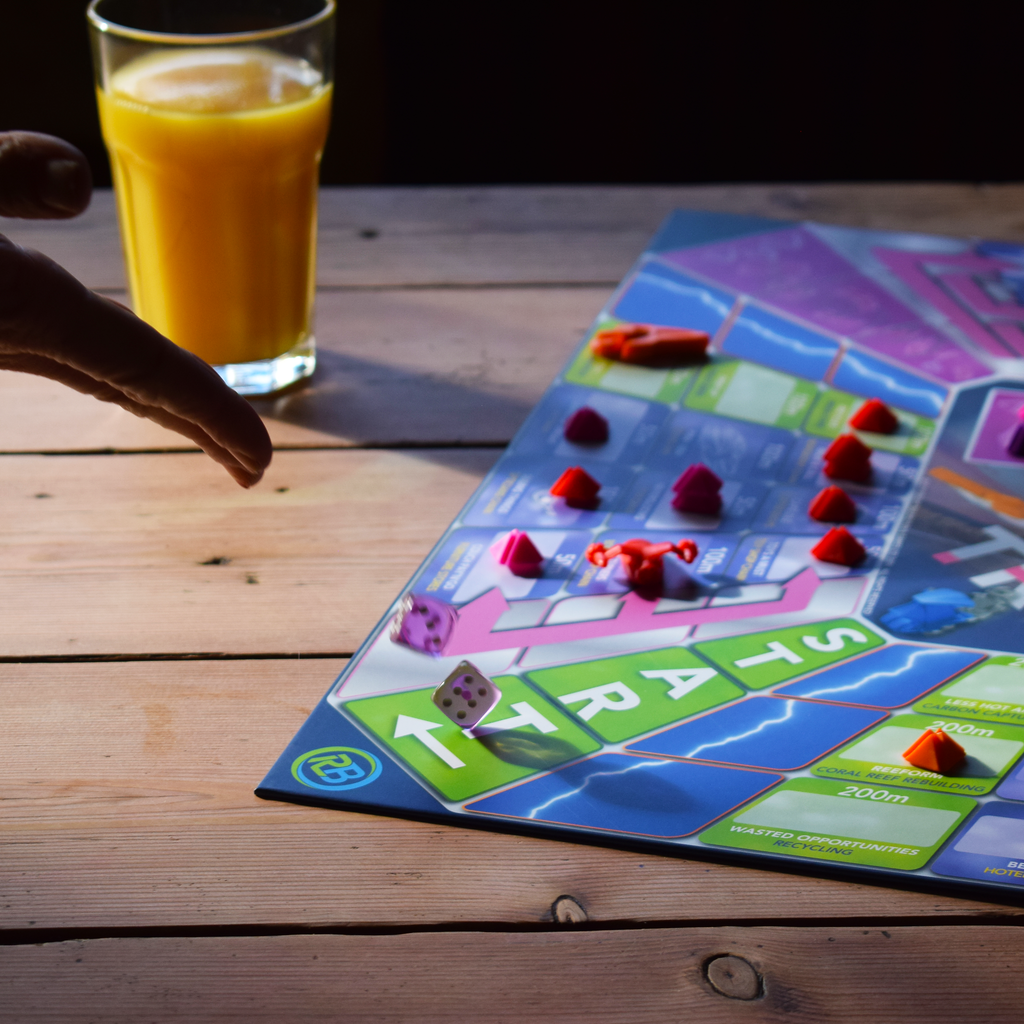 Assets Trip 2–6 player family board game set up on a wooden table, with colourful game pieces and cards spread across the board. Two dice are shown mid-game in the foreground while a player reaches in to take their turn, with a glass of orange juice beside the game. A strategic, replayable board game for family game nights, parties and playing with friends, offering interactive fun for groups.