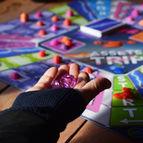 Assets Trip 2–6 player family board game in action, with a player rolling two dice in the foreground while the colourful game board, cards and playing pieces are visible in the background. A strategic and replayable board game, perfect for family game nights, parties and playing with friends. Part of a major board game deal, offering fun and interactive gameplay for groups.