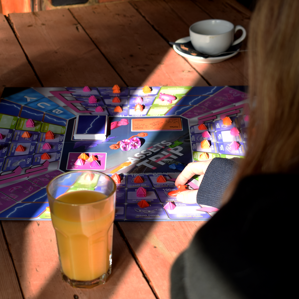 Assets Trip 2–6 player family board game being played at a table, with colourful game pieces, cards and dice spread across the board while players take their turns. A strategic, easy-to-learn and replayable board game for family game nights, parties and playing with friends, offering interactive fun for groups and part of a major board game deal.