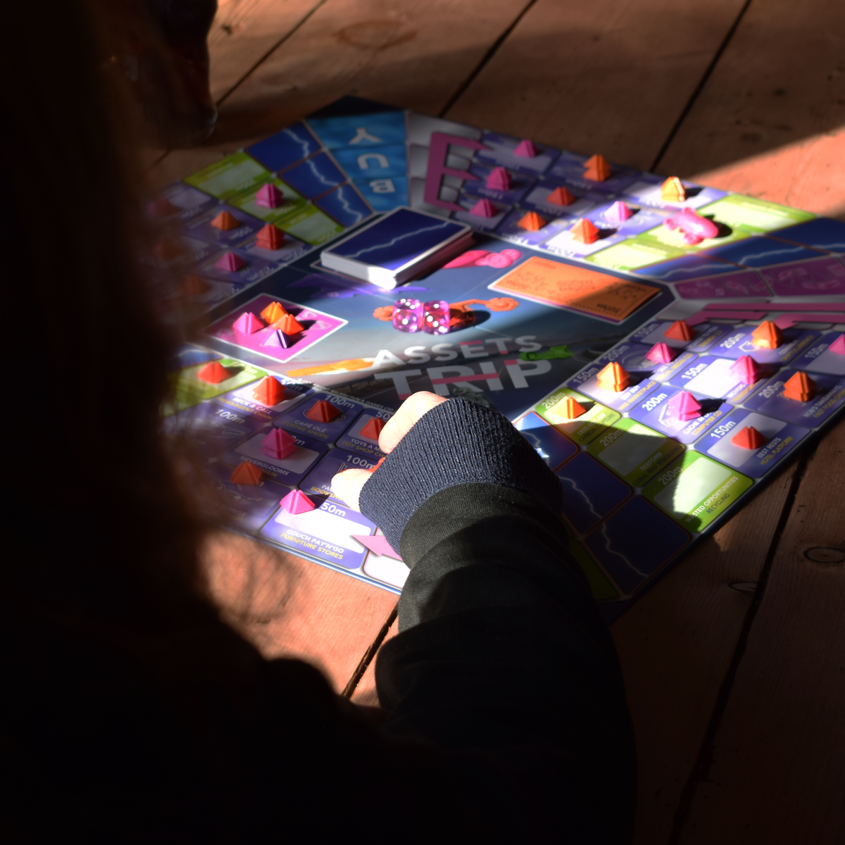 Assets Trip 2–6 player family board game being played outdoors on a wooden table, with colourful game pieces, cards and dice spread across the board while a player reaches in to take their turn. A strategic, easy-to-learn and replayable board game for family game nights, parties and playing with friends, offering interactive fun for groups and part of a major board game deal.