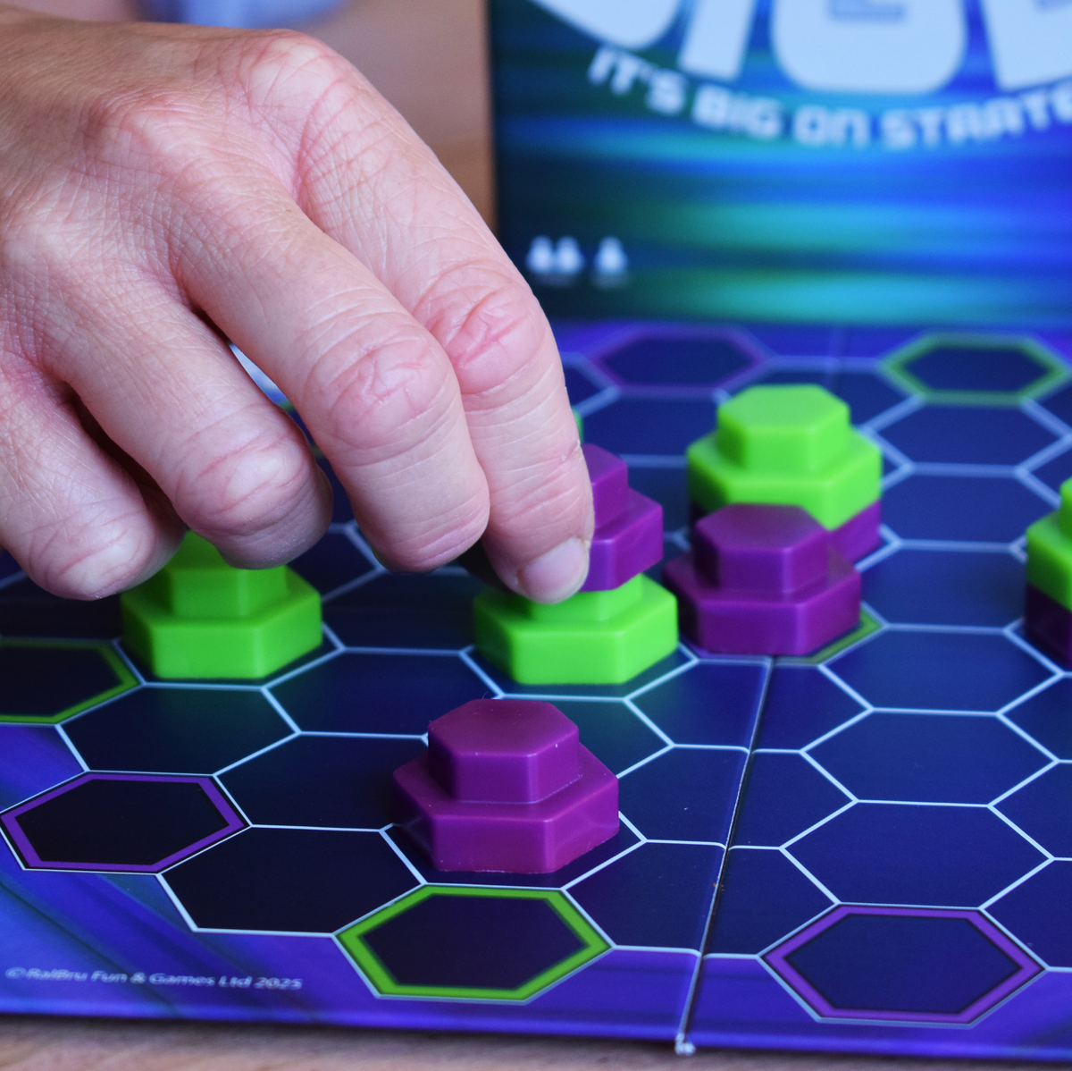 Close-up of a player placing a purple piece on the Bigly hexagonal board, with green and purple pieces in play, highlighting a portable strategy game ideal for travel and game nights.