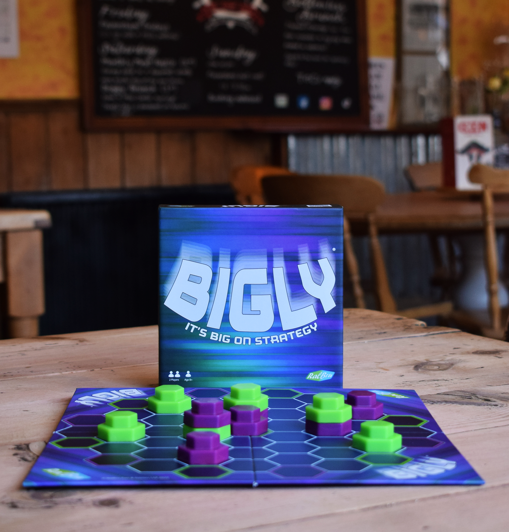 Bigly strategy board game displayed on a wooden café table with the game box and colourful pieces in play, highlighting a replayable two-player game for family and adult game nights.