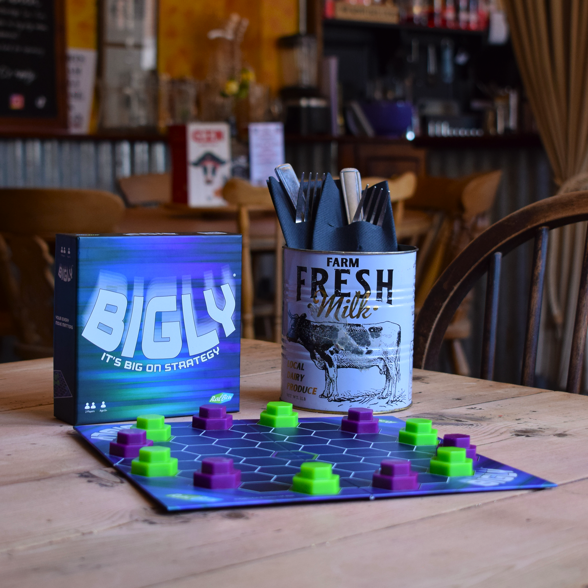 Bigly board game set up on a wooden café table with green and purple playing pieces and the game box displayed behind it, showcasing a travel-friendly abstract strategy game for quick and portable play.