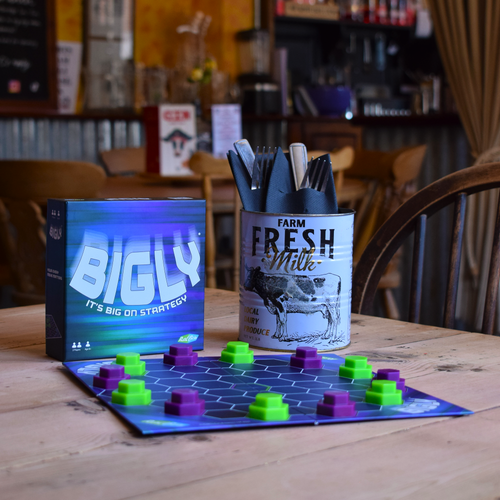 Bigly board game set up on a wooden café table with green and purple playing pieces and the game box displayed behind it, showcasing a travel-friendly abstract strategy game for quick and portable play.