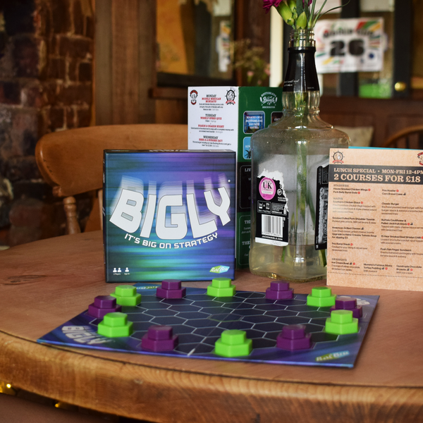 Bigly board game displayed in a relaxed pub setting with the colourful hexagonal game board set up in front of the box, highlighting its compact design, strategic two-player gameplay and portable format for cafés, pubs, holidays and game nights.