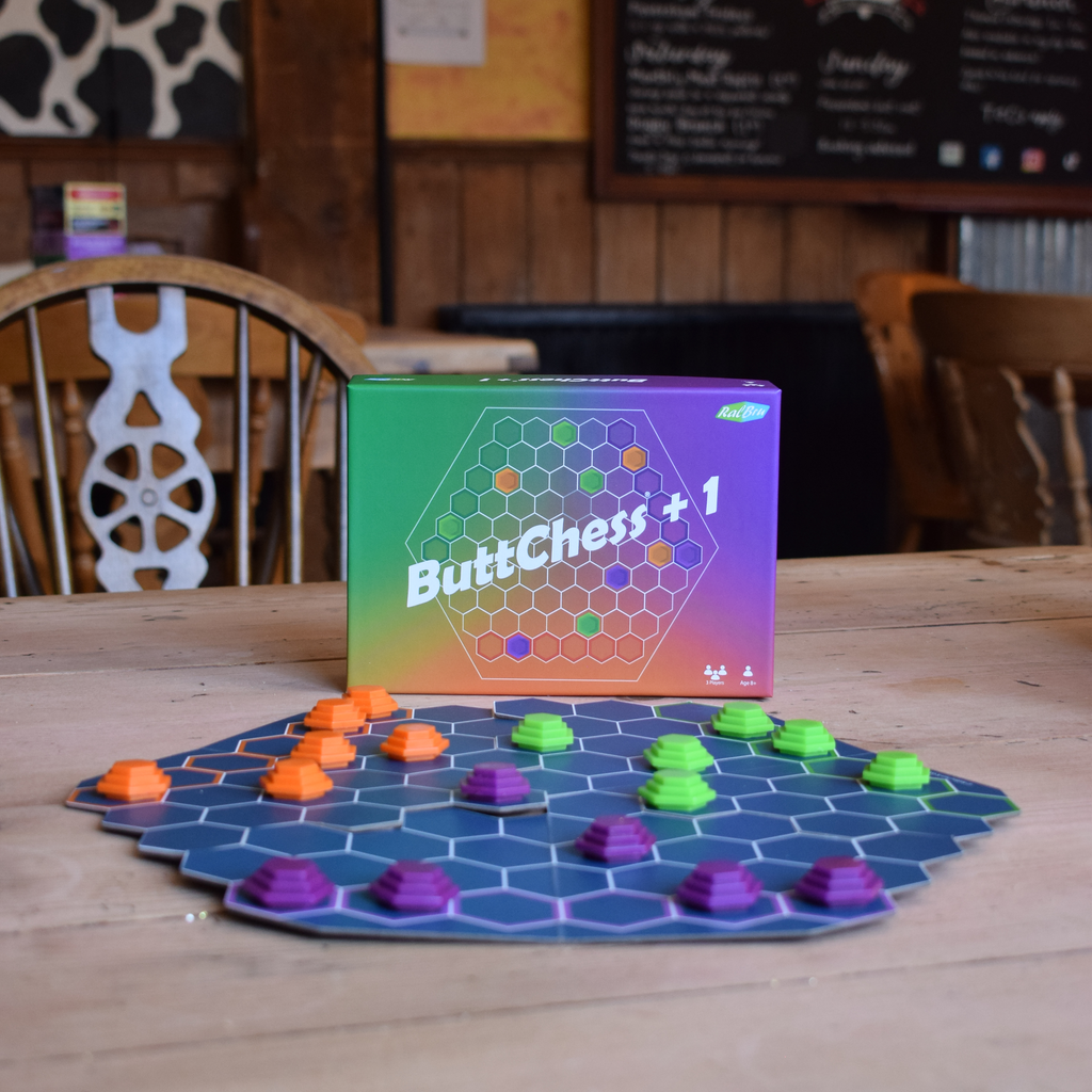 ButtChess + 1 board game set displayed on a wooden table, featuring its colorful gradient box and a blue hexagonal board with orange, green, and purple pieces in a cafe setting.