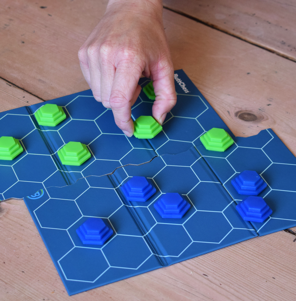 ButtChess travel board game being played, with a player moving a green game piece across the blue hexagonal board. An easy-to-learn, replayable strategy board game for all ages, ideal for family game nights, holidays, travel and playing with friends. An affordable under £20 board game and fun portable travel game.