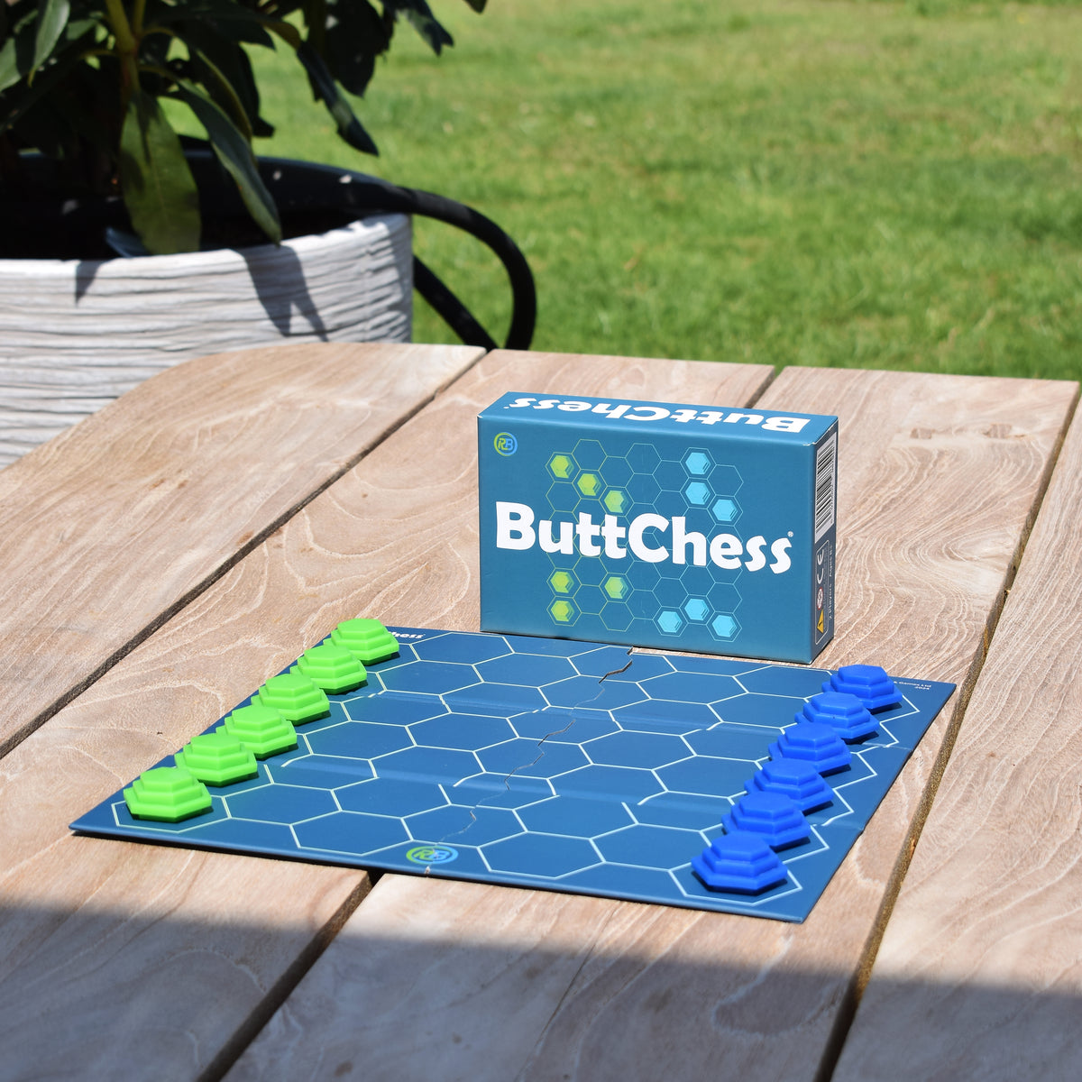 ButtChess travel board game and box displayed outdoors, featuring a portable blue hexagonal game board with green and blue pieces. An easy-to-learn, replayable strategy board game for all ages, perfect for family game nights, holidays, travel and outdoor play. An affordable board game under £20 and a great gift for board game fans.