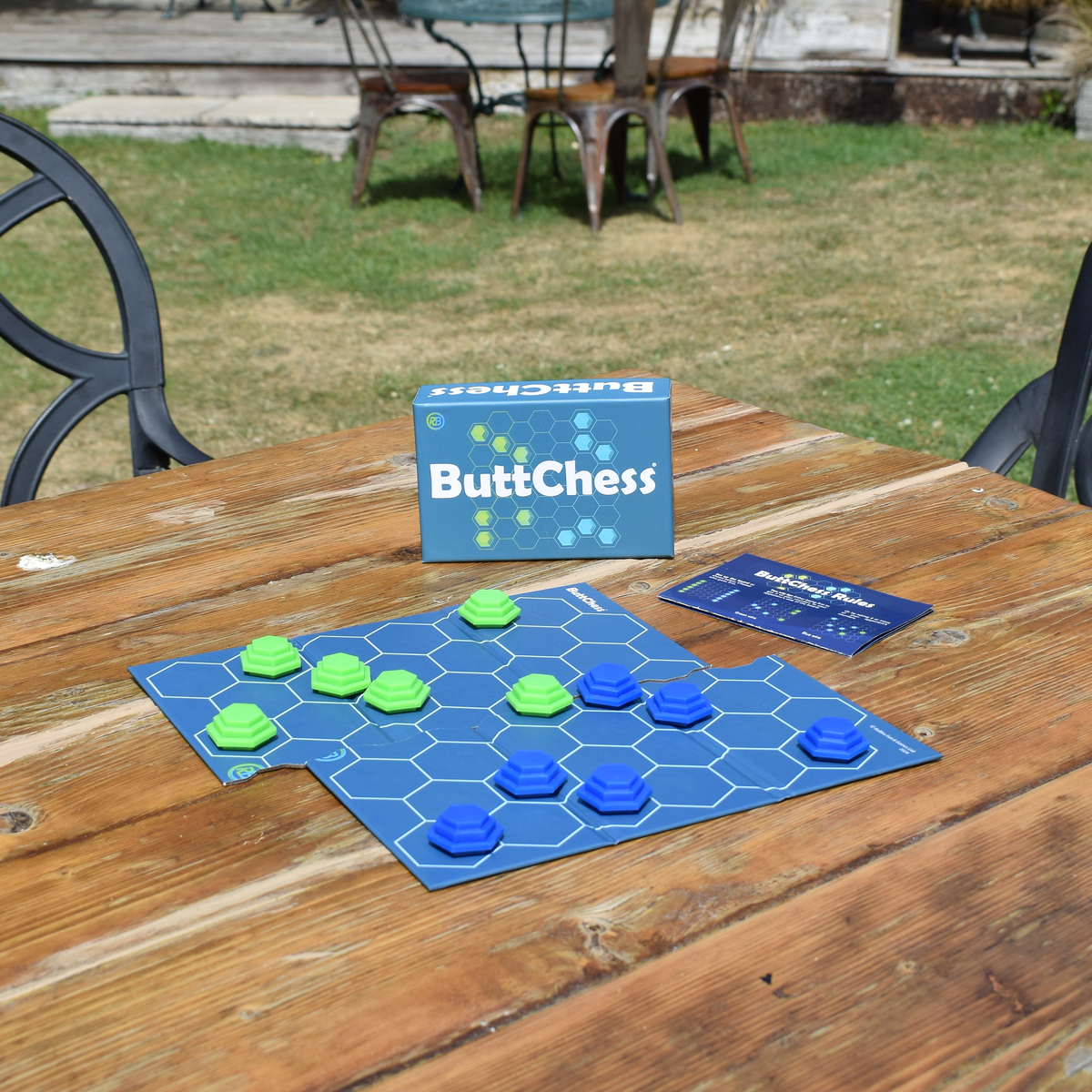 ButChess abstract strategy board game displayed on a wooden outdoor table, with blue and green hexagonal playing pieces, game board, box and rule booklet, showing a compact two-player travel board game for quick strategy play.