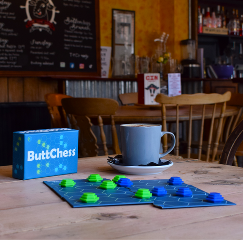A tabletop scene in a cozy cafe or bar interior showing a blue ButtChess game box, a blue chess board with green and blue pieces, a gray mug on a saucer, and wooden chairs in the background.