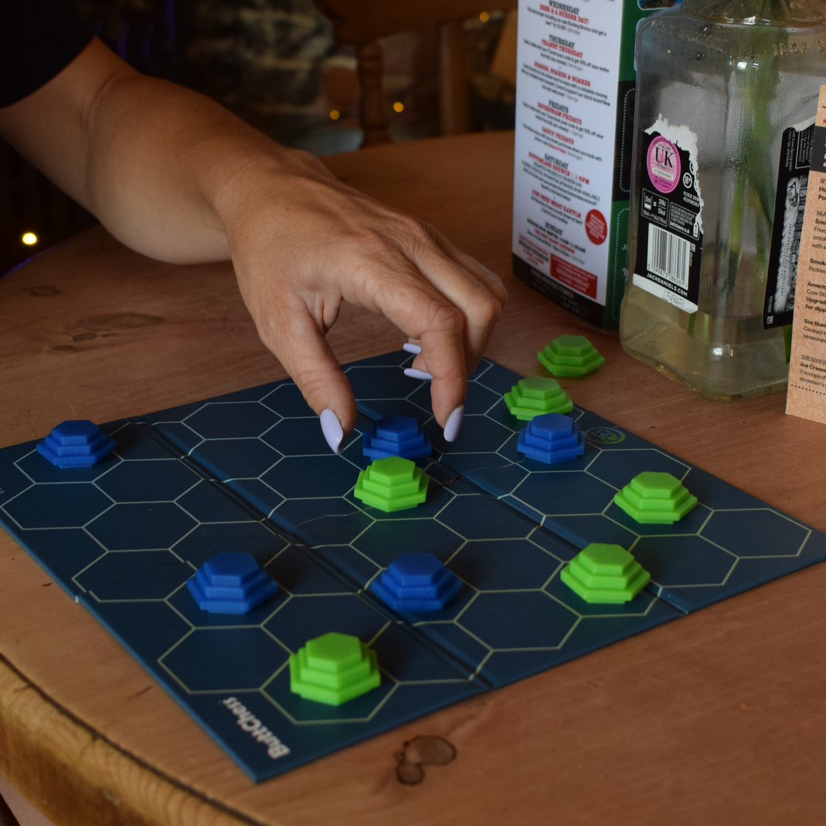ButChess abstract strategy board game being played on a wooden table, with a player moving a blue piece among green and blue hexagonal pieces on the game board, highlighting a quick two-player strategy game for adults.