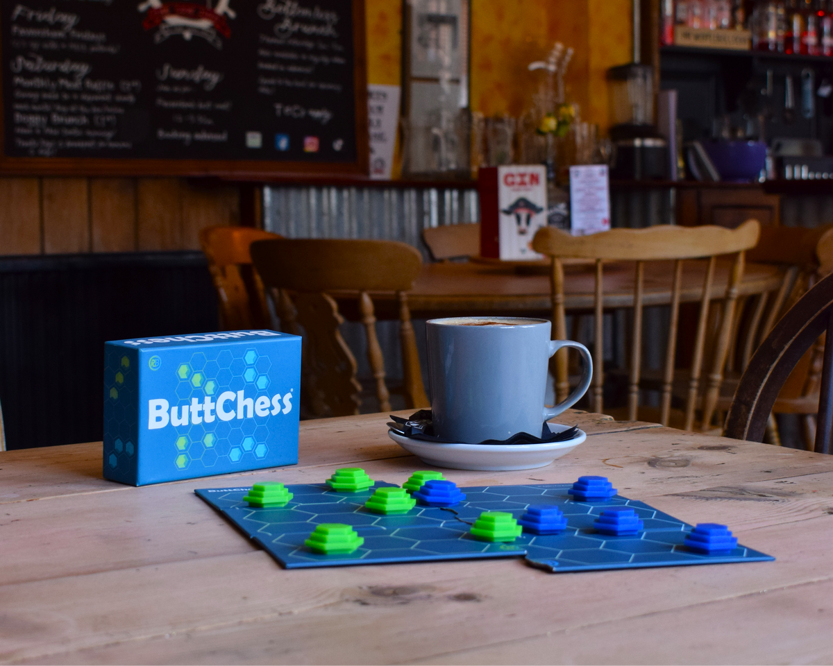 ButChess family board game displayed in a cosy café setting with blue and green playing pieces, game box and coffee cup, showcasing an easy-to-learn, replayable strategy game for all ages.
