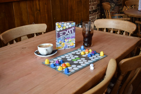 Exit Strategy board game set up on a wooden café table with colourful playing pieces, dice and game box, showcasing a compact travel game priced at £17.99 for affordable strategy play on the go