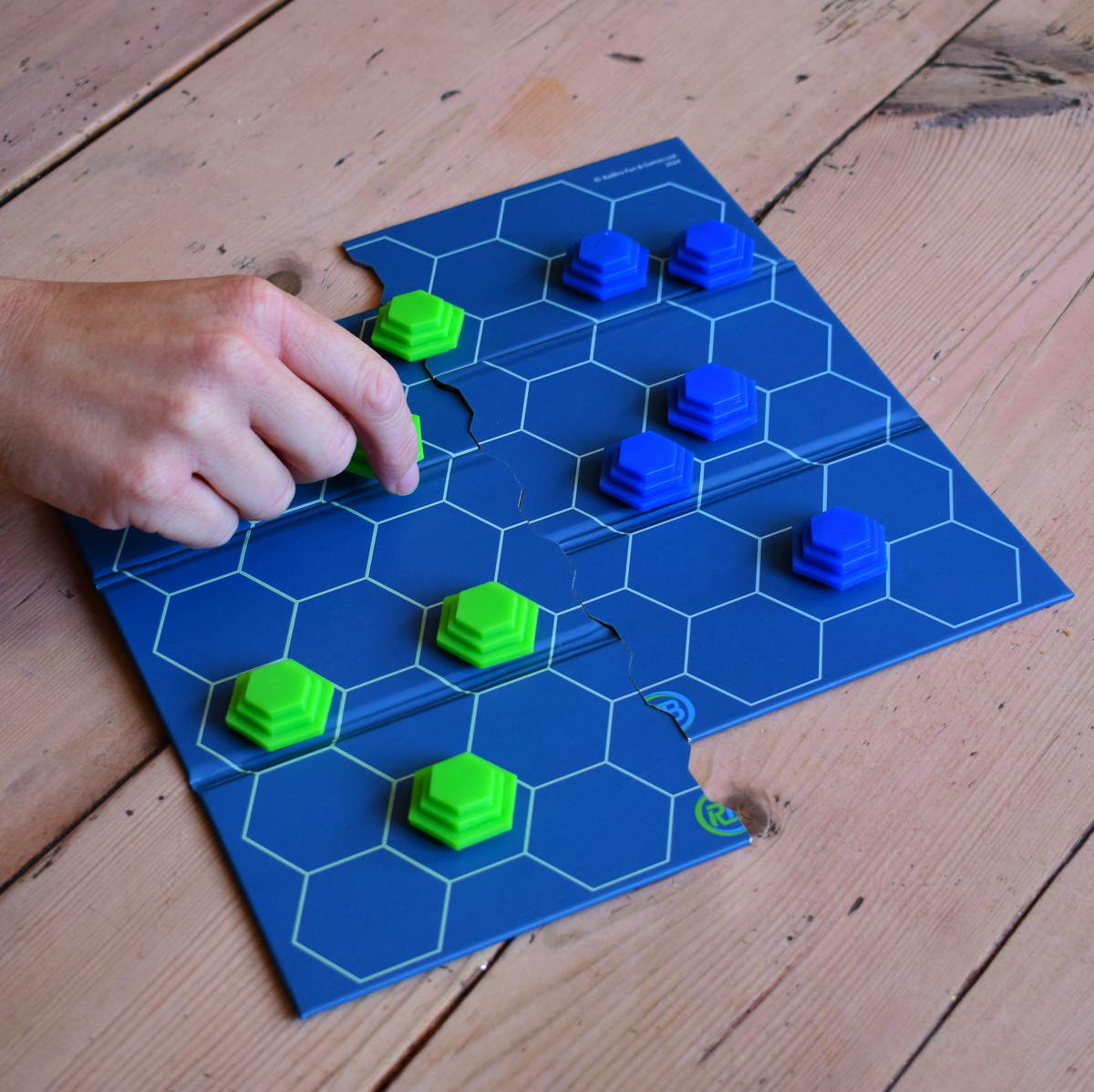 ButtChess travel board game being played, with a player moving a green game piece across the blue hexagonal board. An easy-to-learn, replayable strategy board game for all ages, ideal for family game nights, holidays, travel and playing with friends. An affordable under £20 board game and fun portable travel game.