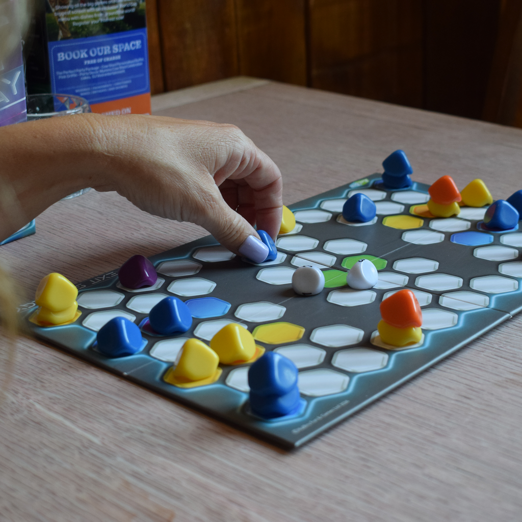 Exit Strategy board game mid-play, showing a player moving a blue counter across the hexagonal board towards the central escape area, with colourful pieces creating a tactical battle. A portable two-player strategy game designed for quick, replayable tabletop gaming.