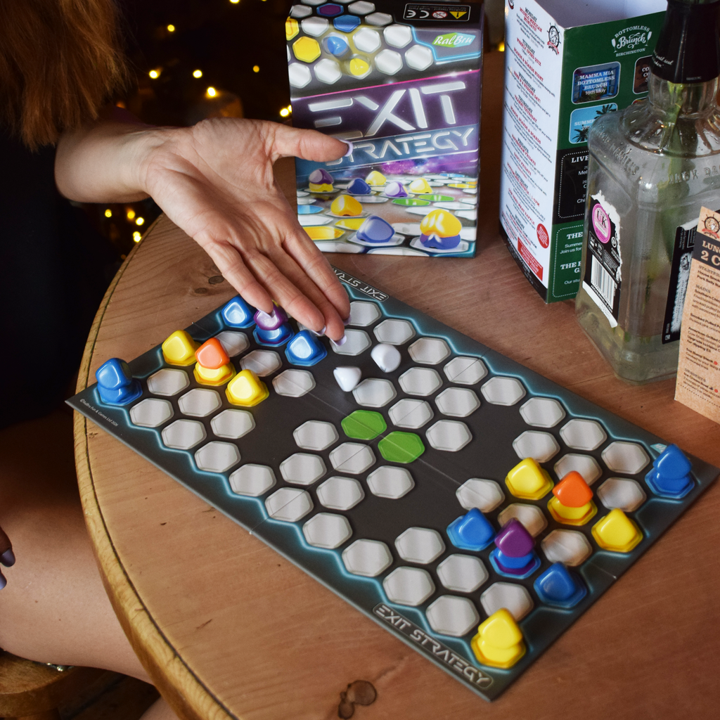 Exit Strategy board game being played at a table, with colourful counters moving across the hexagonal board beside the game box, showcasing tactical two-player gameplay, space-themed abstract strategy and a compact travel board game for quick, replayable play.