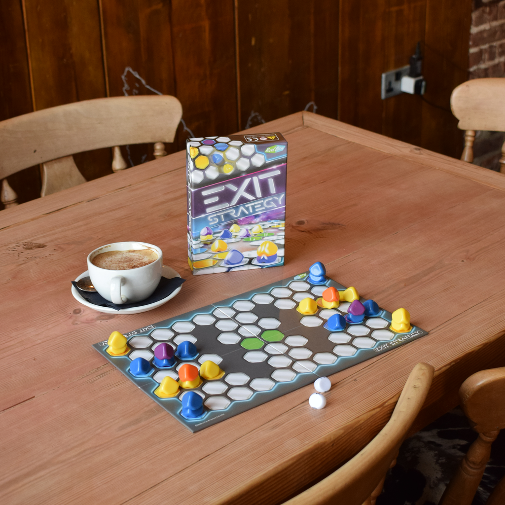 Exit Strategy board game set up on a wooden table beside a coffee, creating a relaxed tabletop gaming scene. The compact space-themed strategy game offers tactical two-player gameplay, portable play and an easy-to-learn challenge for game nights, cafés and travel.