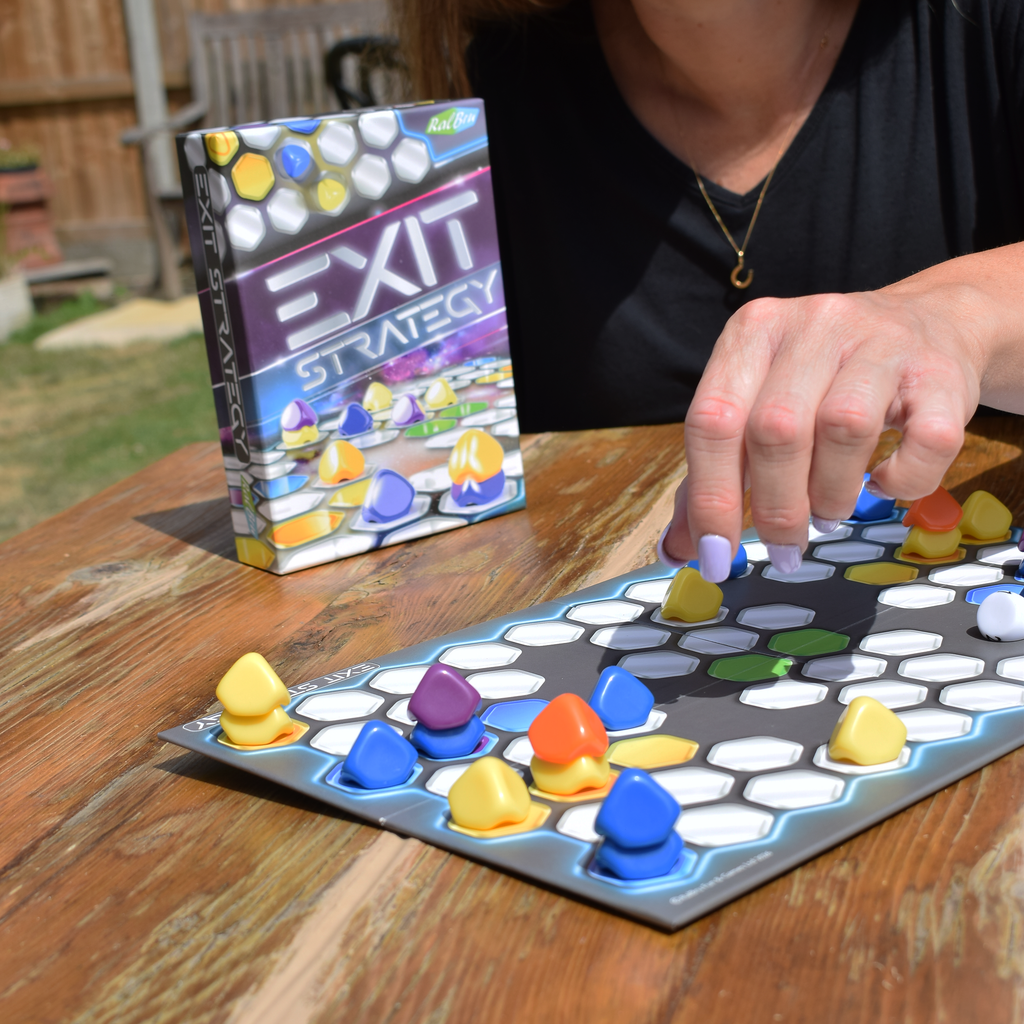 Exit Strategy board game being played outdoors, with a player moving colourful pieces across the hexagonal board beside the game box, highlighting strategic two-player gameplay, space-themed design, portable travel gaming and quick, replayable abstract strategy.